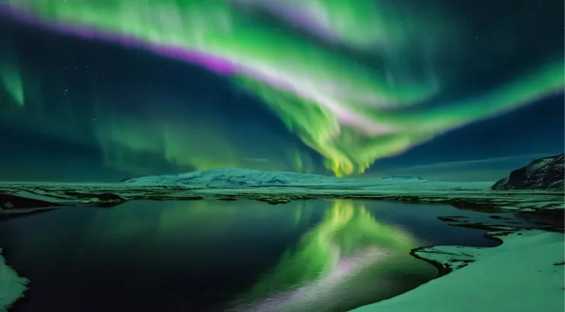 Green aurora reflected in the still water of the glacier lagoon with snow-covered ice in the foreground